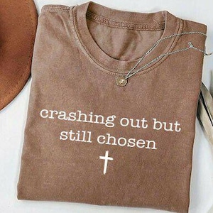 May include: A brown t-shirt with the text "crashing out but still chosen" and a cross symbol. A silver necklace with a small pendant rests on the shirt. A brown hat is visible in the upper left corner.