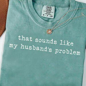 Może przedstawiać: Miętowa koszulka z napisem "that sounds like my husband's problem" wydrukowanym na biało. Złoty naszyjnik z okrągłą zawieszką spoczywa na koszulce. Brązowy kapelusz i suszone liście znajdują się w tle.