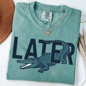 May include: A seafoam green t-shirt with the word "LATER" in navy blue, featuring a navy blue alligator graphic. A gold necklace with a pendant and a brown hat are also visible.