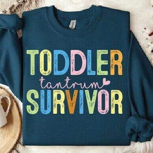 May include: A teal sweatshirt with the words "TODDLER tantrum SURVIVOR" in colorful, block letters. The text is in a playful font, with a small pink heart. The sweatshirt has long sleeves and a crew neck.