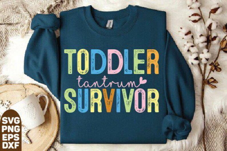 May include: A teal sweatshirt with the words "TODDLER tantrum SURVIVOR" in colorful, block letters. The text is in a playful font, with a small pink heart. The sweatshirt has long sleeves and a crew neck.