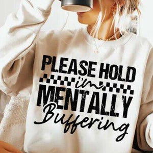 May include: Cream-colored sweatshirt with the text "PLEASE HOLD I'M MENTALLY Buffering" in black. The text is arranged in a bold, eye-catching design. The sweatshirt is a casual, comfortable garment.