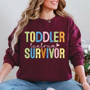May include: A maroon sweatshirt with the words "TODDLER tantrum SURVIVOR" in colorful block letters. The text is in a playful font, with each word in a different color. The sweatshirt is worn by a person.