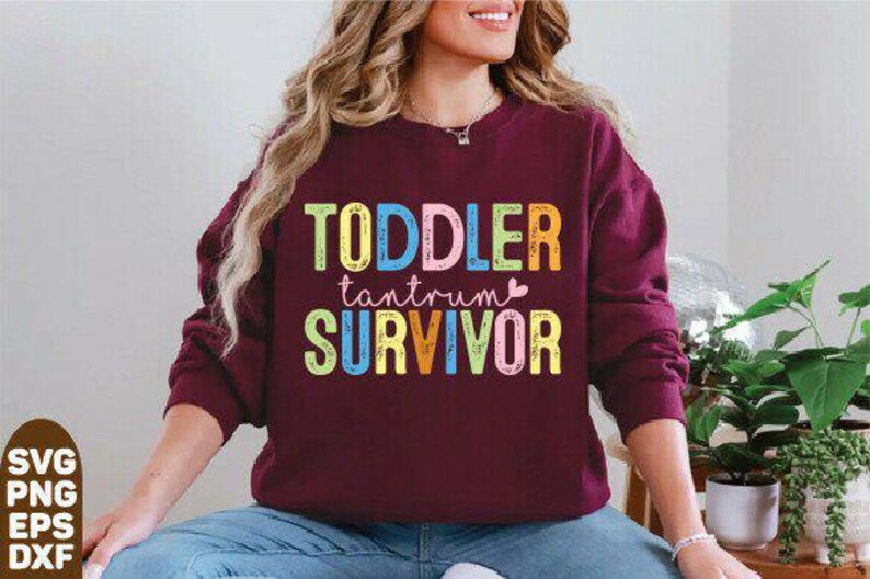 May include: A maroon sweatshirt with the words "TODDLER tantrum SURVIVOR" in colorful block letters. The text is in a playful font, with each word in a different color. The sweatshirt is worn by a person.