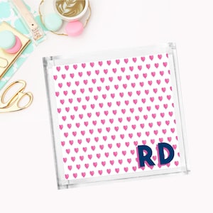 May include: A clear acrylic tray with a pink heart pattern and the initials "RD" in navy blue letters. The tray is perfect for organizing small items on a desk or vanity.