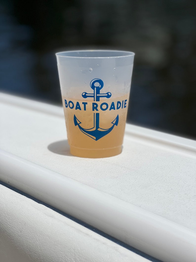 May include: A white plastic cup with a blue anchor and the text "BOAT ROADIE" printed on it. The cup is filled with a light yellow drink.