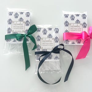 Może przedstawiać: Trzy kalendarze zapakowane w przezroczysty plastik z r&oacute;żnokolorowymi wstążkami. Każdy kalendarz ma wz&oacute;r w kolorze niebieskim i białym z tekstem "kalendarz dwa tysiące dwadzieścia pięć".