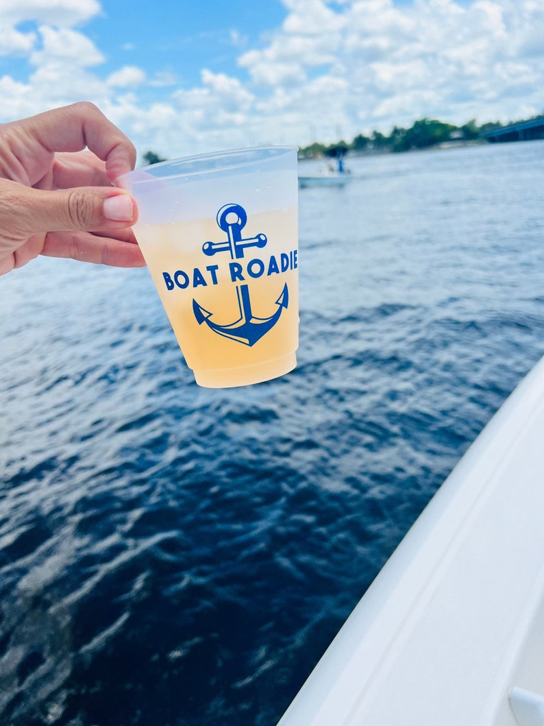 May include: A clear plastic cup with a blue anchor and the text "Boat Roadie" printed on it. The cup is filled with a light yellow beverage. The cup is being held by a hand against a blue water background.