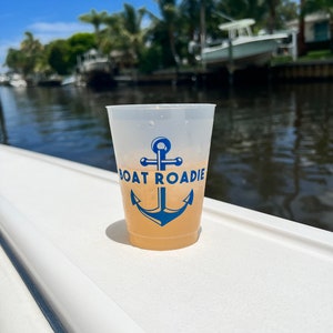 May include: A clear plastic cup with a blue anchor and the text "Boat Roadie" printed on it. The cup is filled with a light yellow beverage and is sitting on a white surface.