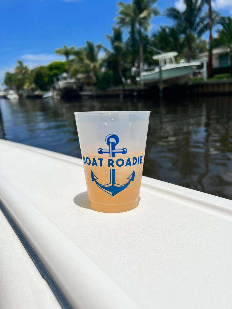 May include: A clear plastic cup with a blue anchor and the text "Boat Roadie" printed on it. The cup is filled with a light yellow beverage and is sitting on a white surface.