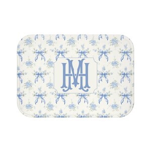 May include: Rectangular bath mat with a floral and bow pattern in light blue and white. The center features a light blue monogram within a white frame. The mat has rounded corners and a soft, plush appearance.