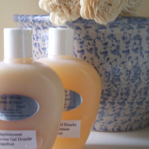 May include: Two clear plastic bottles of shower gel with white caps and labels. The bottles are sitting on a blue and white speckled surface. The label on the left bottle reads "Les Savons, Sels de Nature, Grapefruit, Shower Gel Douche". The label on the right bottle reads "Gel Douche, Mousse, Fruit".