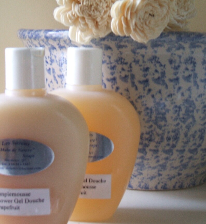 May include: Two clear plastic bottles of shower gel with white caps and labels. The bottles are sitting on a blue and white speckled surface. The label on the left bottle reads "Les Savons, Sels de Nature, Grapefruit, Shower Gel Douche". The label on the right bottle reads "Gel Douche, Mousse, Fruit".