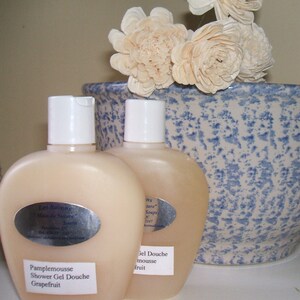 May include: Two clear plastic bottles of shower gel with white caps. The bottles are labeled with the text "Les Savons" and "Main de Nature" in a silver oval. The bottles are sitting on a blue and white ceramic bowl with white flowers.