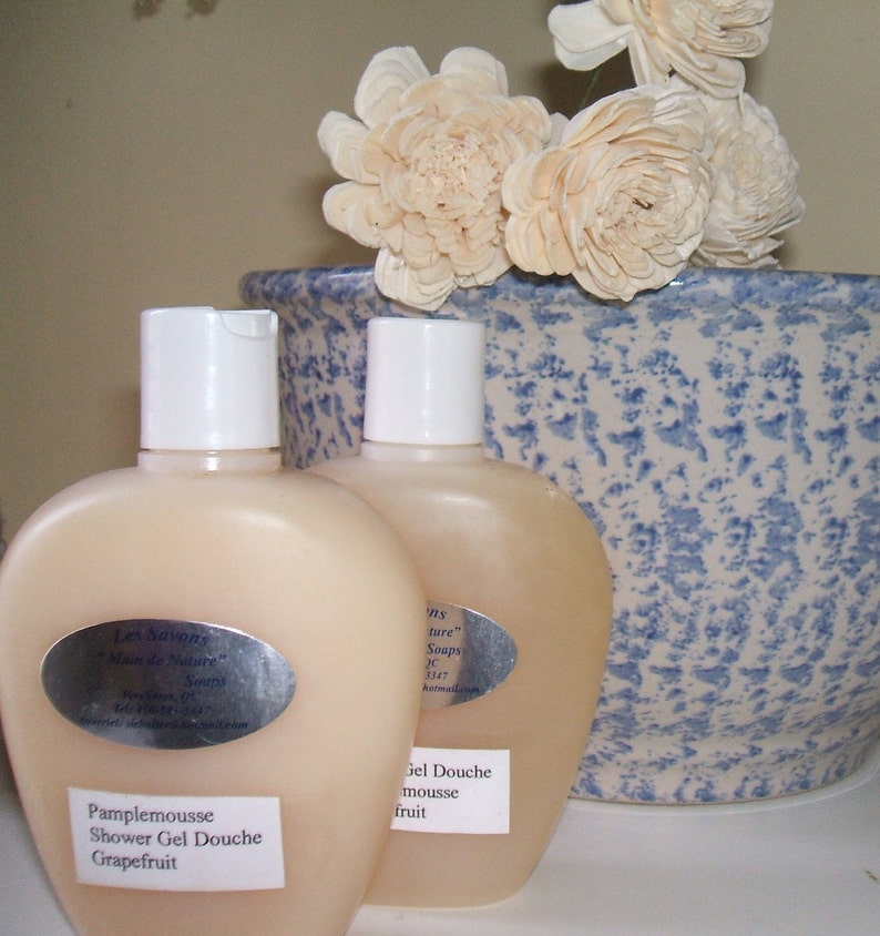 May include: Two clear plastic bottles of shower gel with white caps. The bottles are labeled with the text "Les Savons" and "Main de Nature" in a silver oval. The bottles are sitting on a blue and white ceramic bowl with white flowers.