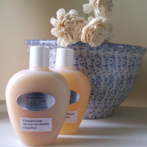 May include: Two clear plastic bottles of shower gel with white caps and labels. The bottle on the left has a label that reads "Pamplemousse Shower Gel Douche Grapefruit". The bottle on the right has a label that reads "Douche use". The bottles are sitting on a white surface in front of a blue and white ceramic bowl with white flowers.