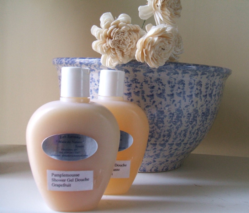 May include: Two clear plastic bottles of shower gel with white caps and labels. The bottle on the left has a label that reads "Pamplemousse Shower Gel Douche Grapefruit". The bottle on the right has a label that reads "Douche use". The bottles are sitting on a white surface in front of a blue and white ceramic bowl with white flowers.