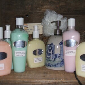 May include: A collection of six bottles of shower gel in various colors and scents, including Pomegranate & Passion, Citrus & Lavender, and Gingerbread. The bottles are arranged on a wooden shelf with a ceramic dispenser in the center.