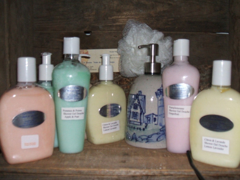 May include: A collection of six bottles of shower gel in various colors and scents, including Pomegranate & Passion, Citrus & Lavender, and Gingerbread. The bottles are arranged on a wooden shelf with a ceramic dispenser in the center.