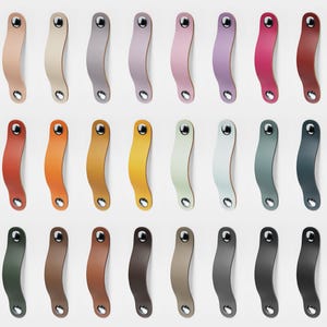 Op de afbeelding: Een verzameling van 27 leren kastgrepen in verschillende kleuren, waaronder wit, beige, grijs, roze, rood, oranje, geel, groen en blauw. Elke greep heeft een gebogen ontwerp met een metalen klinknagel aan elk uiteinde.