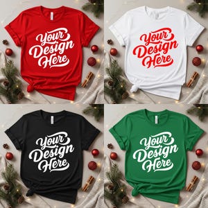 May include: Four t-shirts in red, white, black, and green, each with the text "Your Design Here" in a white, cursive font. The shirts are displayed with holiday decorations, including pine branches, red ornaments, and cinnamon sticks.
