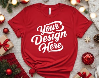 Puede incluir: Una camiseta roja con el texto "Your Design Here" en escritura blanca. La camiseta tiene las mangas enrolladas y un nudo en la parte inferior. La imagen está decorada con adornos navideños y regalos envueltos.
