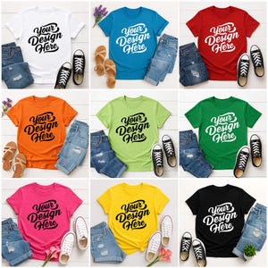 Mockup bundle featuring Gildan 64000 t-shirts in over 70+ colors. The image shows various colored t-shirts with the placeholder text 'Your Design Here' on a flat lay background with flowers, sneakers, and other accessories.