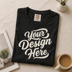 May include: Black t-shirt with the text "Your Design Here" in white script. The shirt has a crew neck and a tag that reads "Comfort Colors". The shirt is folded and placed on a neutral surface.