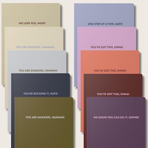 May include: A collection of ten rectangular notebooks in various solid colors including beige, light blue, dark blue, olive green, lavender, pink, coral, brown, and dark purple. Each notebook features personalized text printed on the front cover.