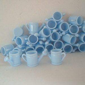 May include: A pile of light blue miniature watering cans. The cans are made of glass and have a spout and handle.