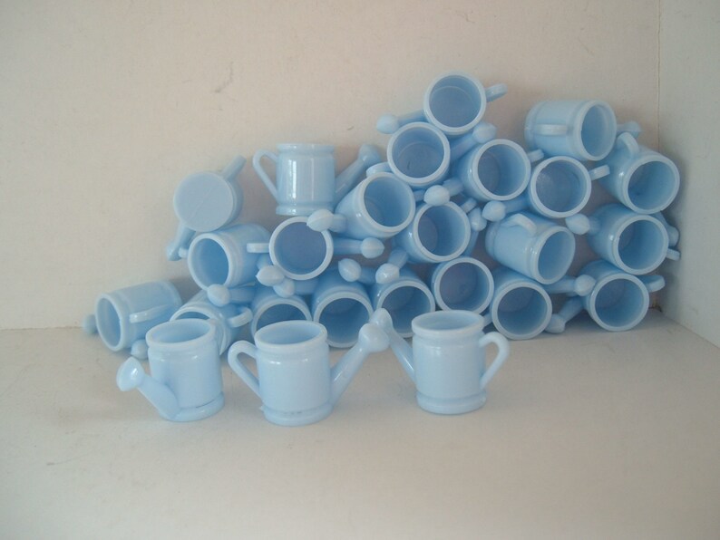 May include: A pile of light blue miniature watering cans. The cans are made of glass and have a spout and handle.