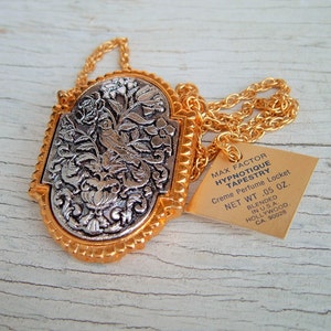 May include: A gold chain necklace with a silver locket featuring an ornate design of a bird and flowers. The locket is attached to a tag that reads "Max Factor Hypnotique Tapestry Creme Perfume Locket Net Wt .05 oz. Blended in U.S.A. Hollywood, CA. 90028"