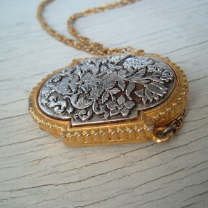 May include: A silver and gold pendant necklace with a floral design. The pendant is oval shaped and has a detailed floral pattern.