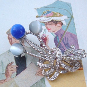 May include: Silver tone brooch with a blue and white stone design. The brooch features a bow with rhinestones and three stones, one blue and two white.