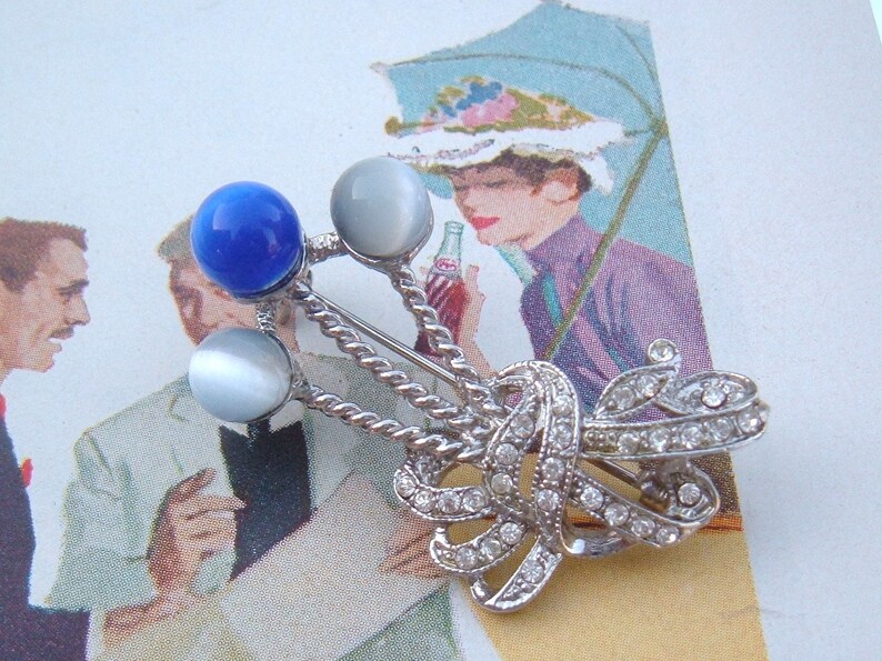 May include: Silver tone brooch with a blue and white stone design. The brooch features a bow with rhinestones and three stones, one blue and two white.