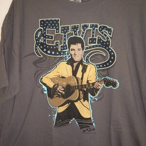 May include: A gray t-shirt with a graphic print of Elvis Presley playing a guitar. The graphic is in blue, yellow, and black. The text "ELVIS" is printed in large letters above the graphic.