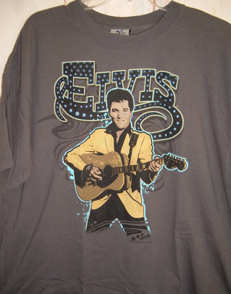 May include: A gray t-shirt with a graphic print of Elvis Presley playing a guitar. The graphic is in blue, yellow, and black. The text "ELVIS" is printed in large letters above the graphic.