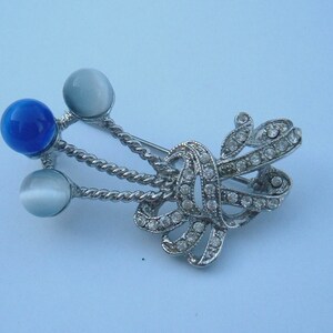 May include: A silver brooch with a blue and white stone design. The brooch features a decorative knot with rhinestones and three round stones.