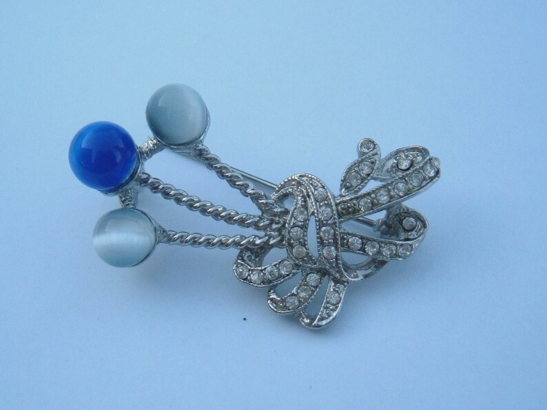 May include: A silver brooch with a blue and white stone design. The brooch features a decorative knot with rhinestones and three round stones.