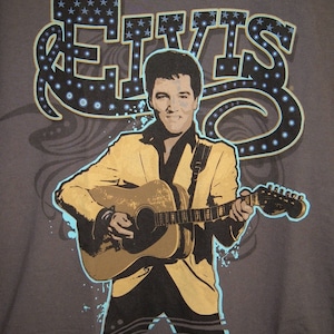 May include: A gray t-shirt with a graphic print of Elvis Presley playing a guitar. The graphic is in a retro style with a blue and white starburst design around the word "Elvis".