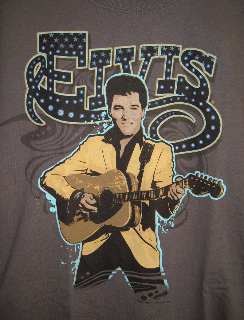 May include: A gray t-shirt with a graphic print of Elvis Presley playing a guitar. The graphic is in a retro style with a blue and white starburst design around the word "Elvis".