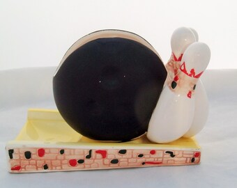 May include: A ceramic bowling pin and ball set. The bowling ball is black with a white rim. The bowling pins are white with red and black stripes. The set is sitting on a yellow and brown base with a brick pattern.