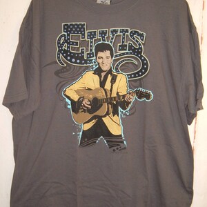 May include: A gray t-shirt with a graphic print of Elvis Presley playing a guitar. The graphic is in a blue, yellow, and black color scheme. The text "ELVIS" is printed in a stylized font above the graphic.