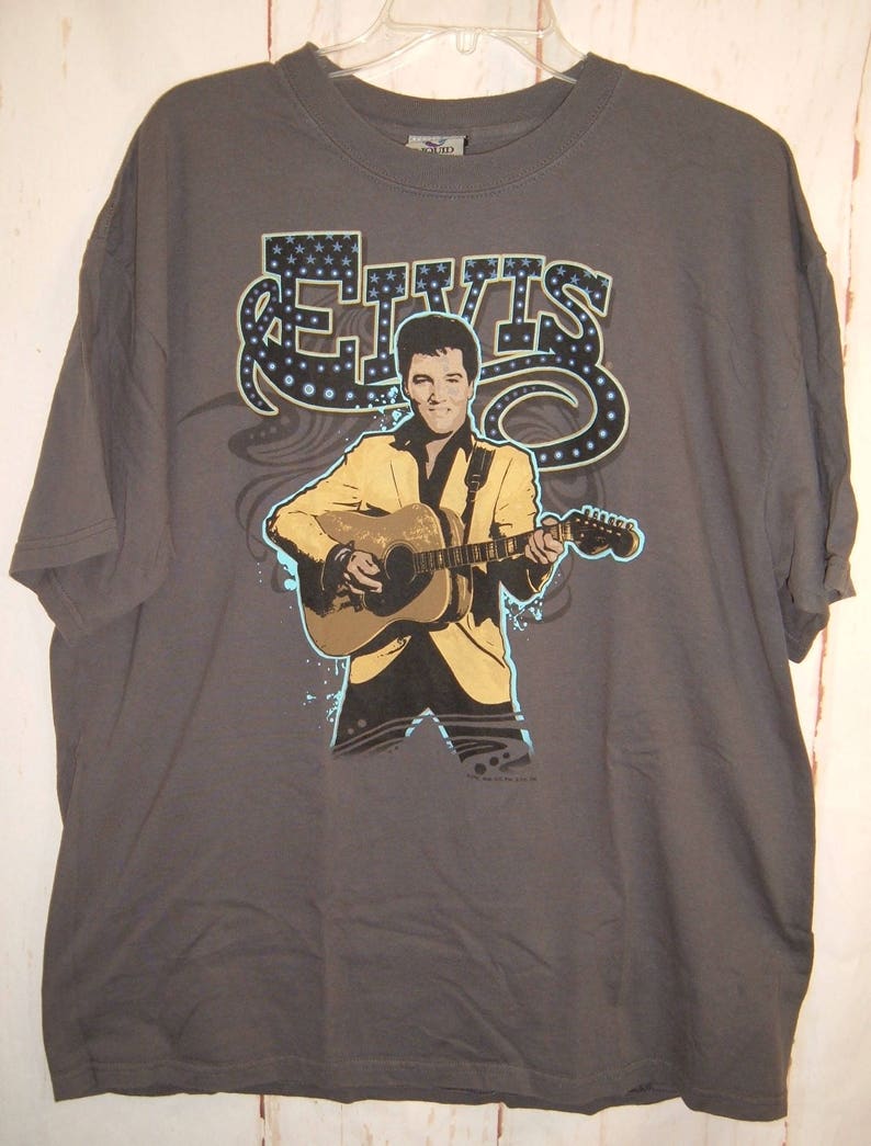 May include: A gray t-shirt with a graphic print of Elvis Presley playing a guitar. The graphic is in a blue, yellow, and black color scheme. The text "ELVIS" is printed in a stylized font above the graphic.