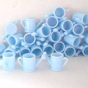 May include: A pile of light blue plastic watering cans, each with a handle and a spout. The watering cans are arranged in a random pattern, with some stacked on top of others.