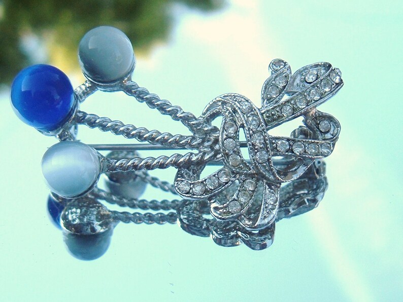 May include: A silver brooch with a bow design and clear rhinestones. The bow is accented with three round stones, two white and one blue.