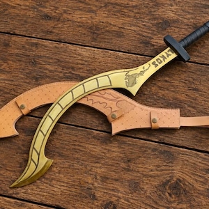 May include: A curved gold-colored sword with a black handle and the word LYKOS engraved on the blade, accompanied by a tan leather sheath featuring a textured surface, metal rivets, and a strap, displayed as a set on a wooden surface.