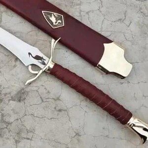以下が含まれることがあります： バーガンディ色の革で包まれた柄と銀色の刃を持つ装飾的な剣。 刃には黒い鳥のデザインが施されています。 剣には、金のアクセントと紋章が付いたバーガンディ色の鞘が付属しています。