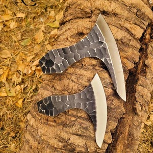 May include: Two small, black, hand-forged axes with hammered steel heads and polished blades. The axes are resting on a piece of tree bark.