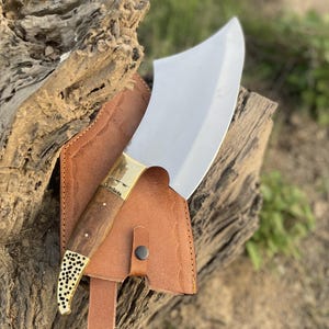May include: A large, stainless steel cleaver with a wooden handle and brass accents, resting in a brown leather sheath. The cleaver has a curved blade and is designed for chopping. The handle is dark brown with a decorative brass end.